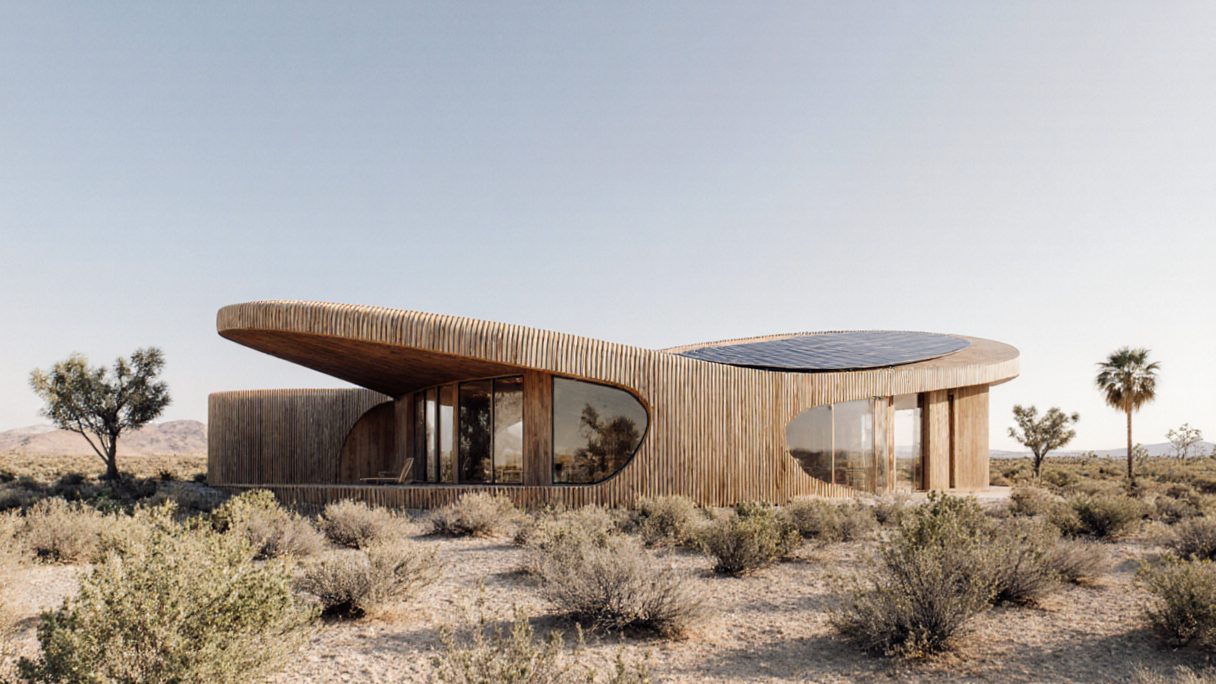 A low desert house clad in vertical timber battens with curved roof planes, rooftop solar panels and Joshua trees under a pale sky