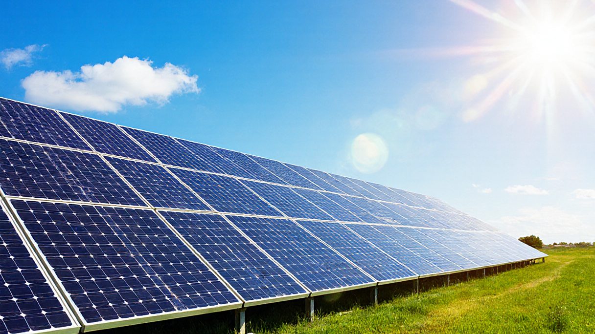 A long row of photovoltaic panels angled to a bright sun over open grassland