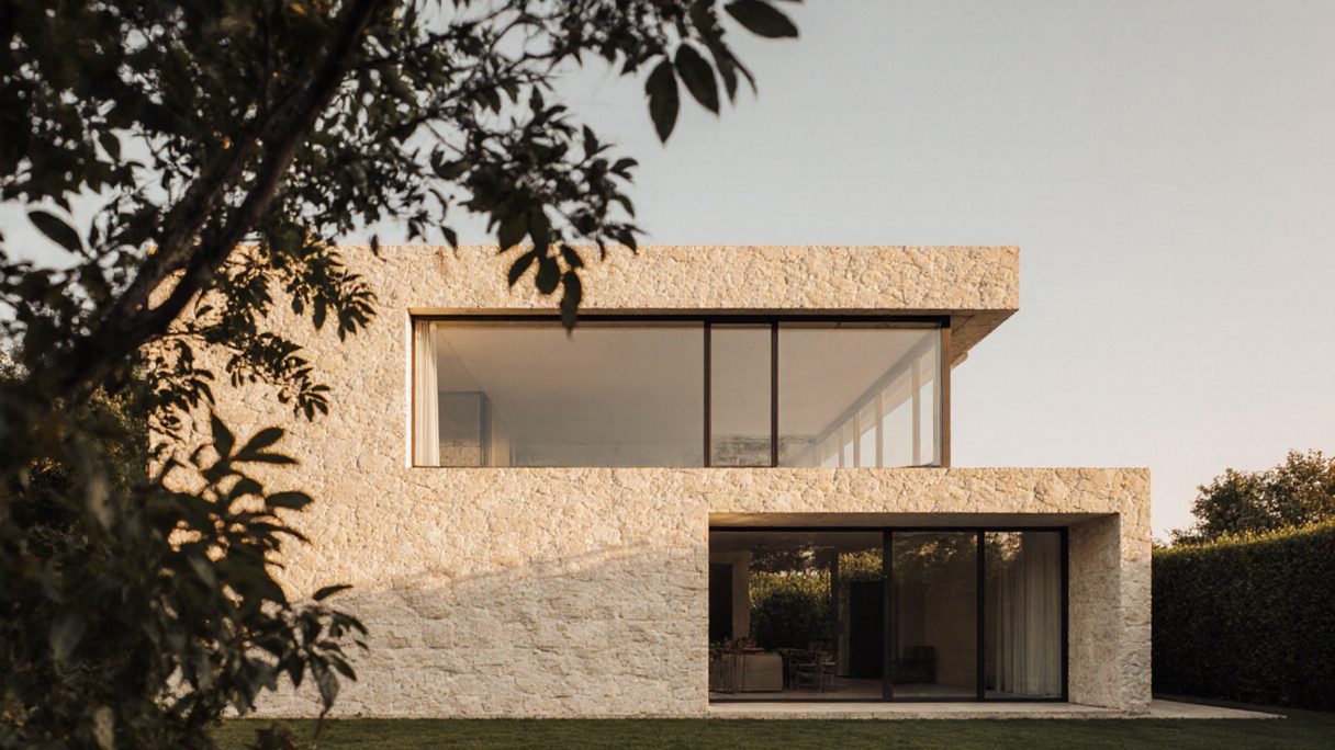 A stone-clad house with stacked terraces and full-height glazing, shaded by a mature tree