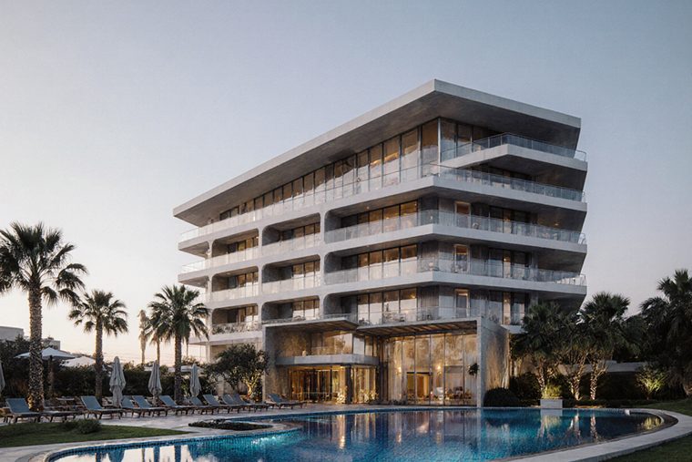 A low-rise hotel with stacked balconies and date palms, lit warm at dusk beside a pool deck