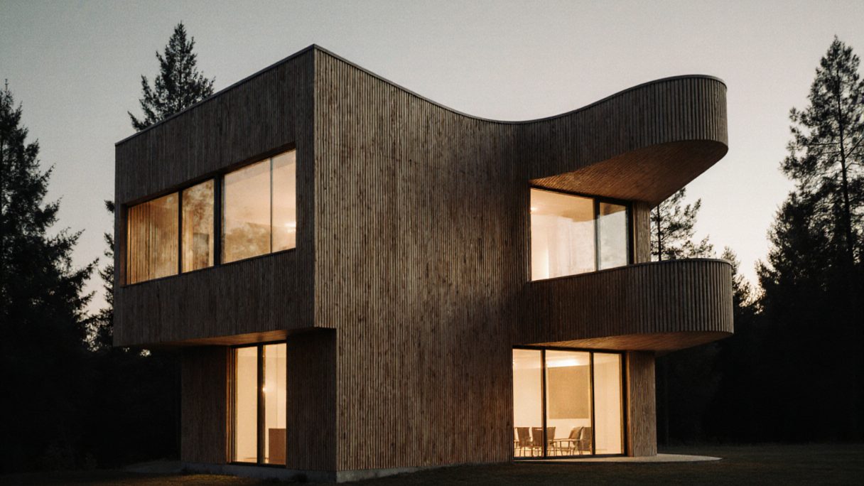 A dark timber house at dusk with a curved balcony, every window lit from inside